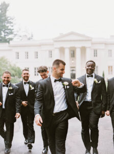 A group of groomsmen walk together in tuxedos in front of a grand white estate, adding a lively and celebratory moment to a Wadsworth Mansion wedding day in Connecticut.
