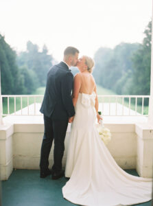 The bride and groom share a quiet kiss on a balcony overlooking expansive green lawns, capturing the serene setting of a Wadsworth Mansion CT wedding.