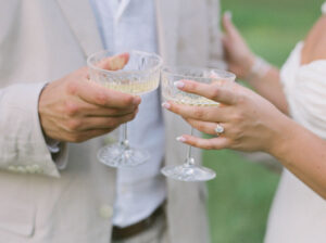 The Chanler at Cliff Walk Wedding | Deirdre Photography Timeless and Organic Moments | Two people clink crystal champagne glasses outdoors. One wears a light suit, the other a white dress with an elegant ring, suggesting a celebratory moment.