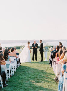 Eisenhower House Wedding | Deirdre Photography Timeless and Organic Moments | A bride and groom stand before an officiant on a grassy field during an outdoor wedding. Guests sit on white chairs, with a serene lake backdrop.