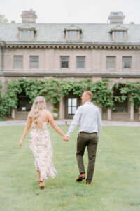 Harkness Park Wedding | Deirdre Photography Timeless and Organic Moments | A couple holding hands walks across a grassy lawn towards a large, ivy-covered building. The scene conveys romance and tranquility on a clear day.