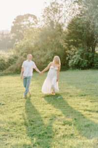 Harkness Park Wedding | Deirdre Photography Timeless and Organic Moments | A couple walks hand in hand on a sunlit grassy field, surrounded by lush trees. The scene conveys warmth and serenity, evoking a sense of romance.