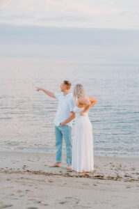 Harkness Park Wedding | Deirdre Photography Timeless and Organic Moments | A couple stands on a beach at sunset, both wearing white. The man points toward the ocean, creating a serene and romantic atmosphere.