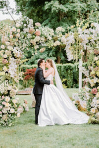 Bride and groom kissing beneath a floral-covered ceremony arbor surrounded by lush garden blooms at one of the most romantic Newport wedding venues.