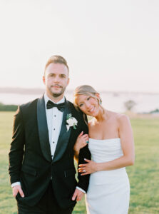 Eisenhower House Wedding | Deirdre Photography Timeless and Organic Moments | A couple poses in wedding attire on a sunlit field. The groom wears a black tuxedo and bow tie; the bride, in a strapless white gown, leans affectionately on him.