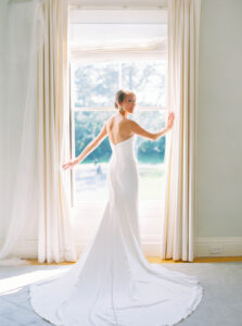 Eisenhower House Wedding | Deirdre Photography Timeless and Organic Moments | Bride in a long white gown stands by a bright window, back towards the camera, with a serene expression. Curtains frame the sunlight streaming in.