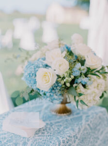 The Chanler at Cliff Walk Wedding | Deirdre Photography Timeless and Organic Moments | A delicate floral arrangement with white roses and blue hydrangeas sits on a lace-draped table. The setting is outdoors, conveying a serene, elegant atmosphere.