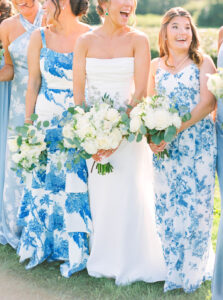 The Chanler at Cliff Walk Wedding | Deirdre Photography Timeless and Organic Moments | Three women in floral dresses and a white gown smile brightly, holding white bouquets. The setting is outdoors, with a cheerful and joyful vibe.