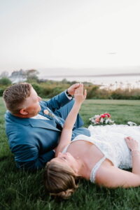 Wedding Photographers Newport RI | Deirdre Photography Timeless and Organic Moments | A couple in wedding attire reclines on grass, holding hands and gazing at the sky. A scenic backdrop of distant water