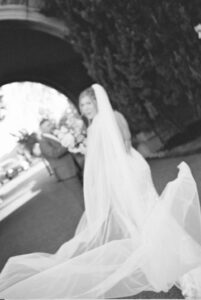 Wedding Photographers Newport RI | Deirdre Photography Timeless and Organic Moments | A black and white photo shows a bride in a flowing dress and veil standing under an archway. A blurred groom holds flowers in the background.