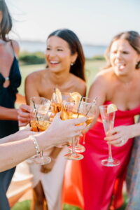 Wedding Photographers Newport RI | Deirdre Photography Timeless and Organic Moments | Smiling women in bright dresses raising glasses for a toast outdoors. The scene conveys joy and celebration.