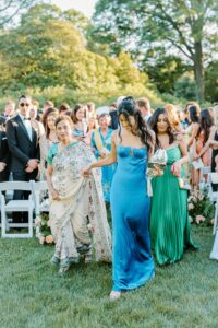 Brooklyn Botanic Garden Wedding | Deirdre Photography Timeless and Organic Moments | A woman in a blue dress and another in green walk an elderly woman in a sari down a grassy aisle, surrounded by guests in formal attire.