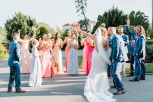 Wedding Photographers Newport RI | Deirdre Photography Timeless and Organic Moments | A bridal party celebrates outdoors, featuring bridesmaids in pink dresses and groomsmen in blue suits. The bride holds a bouquet, exuding joy and festivity.
