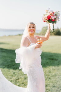 Wedding Photographers Newport RI | Deirdre Photography Timeless and Organic Moments | A joyful bride in a white gown holds a vibrant bouquet and glass, smiling widely. She stands on a lush lawn with water and trees in the background.