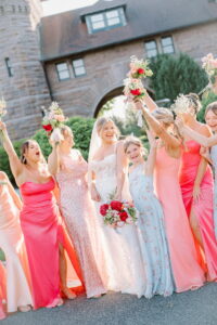 Wedding Photographers Newport RI | Deirdre Photography Timeless and Organic Moments | Bride in white lace gown and veil, surrounded by joyful bridesmaids in pink and floral dresses, holding bouquets aloft. Stone building in background.