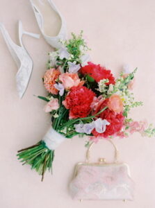 Wedding Photographers Newport RI | Deirdre Photography Timeless and Organic Moments | A vibrant bridal bouquet of red, pink, and peach flowers rests on a soft pink background beside elegant white lace heels and a delicate floral clutch.