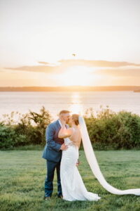 Wedding Photographers Newport RI | Deirdre Photography Timeless and Organic Moments | A couple in wedding attire embraces and kisses on a green lawn, backlit by a sunset over a serene body of water.