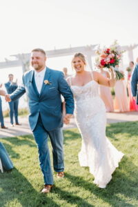 Wedding Photographers Newport RI | Deirdre Photography Timeless and Organic Moments | A joyful couple walks hand in hand outdoors, the bride in a white gown holding a vibrant bouquet, the groom in a blue suit.