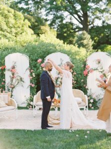 Brooklyn Botanic Garden Wedding | Deirdre Photography Timeless and Organic Moments | A couple in wedding attire exchanges garlands in a lush garden setting. Vibrant flowers decorate white panels.