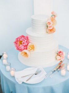 The Chanler at Cliff Walk Wedding | Deirdre Photography Timeless and Organic Moments | A three-tier white wedding cake with pink and peach flowers is on a blue table. Surrounded by candles, it conveys elegance and romance.