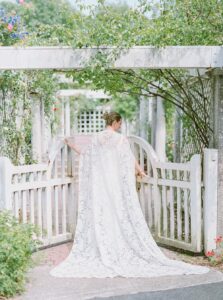 Brooklyn Botanic Garden Wedding | Deirdre Photography Timeless and Organic Moments | A bride in a delicate, lace gown stands facing a white garden gate, surrounded by lush greenery and roses.