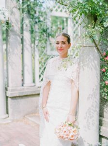 Brooklyn Botanic Garden Wedding | Deirdre Photography Timeless and Organic Moments | A woman in a white lace dress stands against a stone pillar in a garden, holding a bouquet of pink roses.