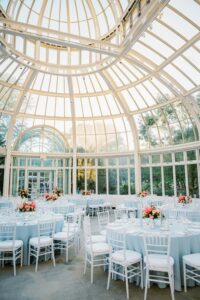 Brooklyn Botanic Garden Wedding | Deirdre Photography Timeless and Organic Moments | Elegant glass-walled venue with a domed ceiling. Tables draped in light blue, adorned with vibrant floral centerpieces. Sunlight filters through, creating a serene ambiance.