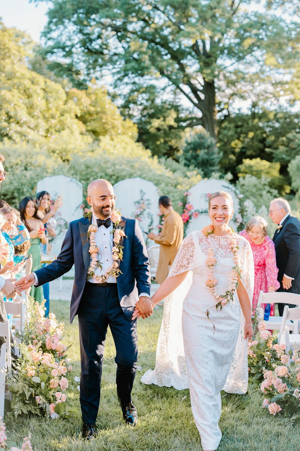 Brooklyn Botanic Garden Wedding | Deirdre Photography Timeless and Organic Moments | A joyful newlywed couple walks hand in hand down an aisle adorned with flowers. Both wear elegant attire and floral garlands, with happy guests in the background.