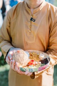 Brooklyn Botanic Garden Wedding | Deirdre Photography Timeless and Organic Moments | A person in a tan traditional outfit holds a metal tray with a coconut, spices, incense, and small bowls in sunlight