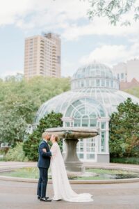 Brooklyn Botanic Garden Wedding | Deirdre Photography Timeless and Organic Moments | A couple sharing a kiss in front of a glass-domed conservatory. The bride wears a flowing white gown, the groom is in a blue suit. Lush greenery and city skyline in the background.