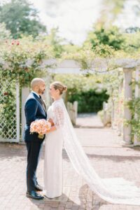 Brooklyn Botanic Garden Wedding | Deirdre Photography Timeless and Organic Moments | A bride in a lace gown and long train holds peach roses, standing with a groom in a dark suit. They face each other in a sunlit garden path.