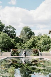 Brooklyn Botanic Garden Wedding | Deirdre Photography Timeless and Organic Moments | A couple stands at the edge of a serene, reflective pond, surrounded by lush greenery and steps.