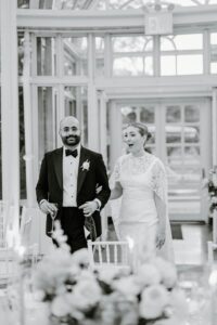 Brooklyn Botanic Garden Wedding | Deirdre Photography Timeless and Organic Moments | A couple walks arm in arm at their wedding reception. The man wears a tuxedo, and the woman, in a lace bridal gown, looks surprised. The setting is elegant, with white floral decorations and soft lighting, conveying joy and celebration.