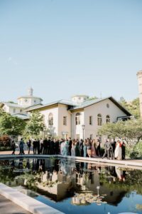 Brooklyn Botanic Garden Wedding | Deirdre Photography Timeless and Organic Moments | A joyful wedding crowd gathers by a reflective pond, with lily pads, near an elegant, light-colored building under a clear blue sky.