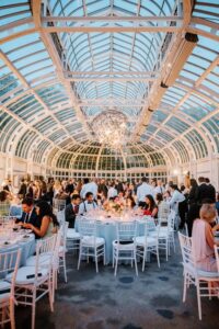 Brooklyn Botanic Garden Wedding | Deirdre Photography Timeless and Organic Moments | Elegant event in a glass-ceilinged conservatory, featuring round tables with white chairs, guests in formal attire, and a central chandelier.