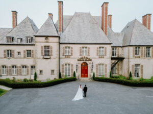 Newport Rhode Island Wedding Photographers | Deirdre Photography Timeless and Organic Moments | A bride in a white gown and a groom in a black suit stand before an elegant mansion with multiple chimneys and a grand red door