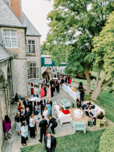 Newport Rhode Island Wedding Photographers | Deirdre Photography Timeless and Organic Moments | Elegant outdoor party at a large stone house, featuring guests in formal attire, a buffet table, and lounge seating under lush trees, creating a lively atmosphere.