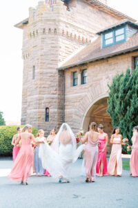 Wedding Photographers Newport RI | Deirdre Photography Timeless and Organic Moments | Bride with veil walks outside a stone building, surrounded by bridesmaids in pastel dresses holding flowers.