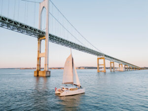 Wedding Photographer Newport Rhode Island | Deirdre Photography Timeless and Organic Moments | A sailboat glides on calm water beneath a large suspension bridge at sunset. The sky is clear, and the scene conveys tranquility