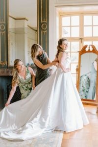 Newport Rhode Island Wedding Photographers | Deirdre Photography Timeless and Organic Moments | Bride in a flowing white dress stands in a bright room with two friends helping adjust her gown. A mirror reflects the scene, adding warmth and anticipation.