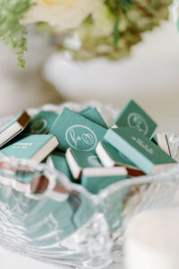 Personalized green matchboxes displayed in a glass tray alongside floral details and soft editorial styling at a Newport Rhode Island wedding venue.