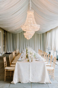 Newport Rhode Island Wedding Photographers | Deirdre Photography Timeless and Organic Moments | Elegant dining setup under a large, ornate chandelier in a tented space. Long table with white cloth, golden chairs, candles, and floral centerpieces