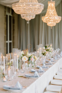 Newport Rhode Island Wedding Photographers | Deirdre Photography Timeless and Organic Moments | Elegant dining table set for a wedding, adorned with white flowers, glassware, and folded napkins. Crystal chandeliers and candles add a warm glow.