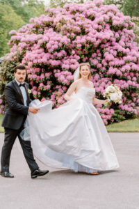 Newport Rhode Island Wedding Photographers | Deirdre Photography Timeless and Organic Moments | A joyful bride in a white gown is helped by a suited man to carry her train. They pose near a blooming pink rhododendron bush