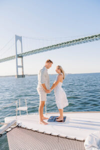 Wedding Photographer Newport Rhode Island | Deirdre Photography Timeless and Organic Moments | A couple holds hands on a boat deck, smiling at each other under a clear blue sky. A large suspension bridge spans the background over the water.