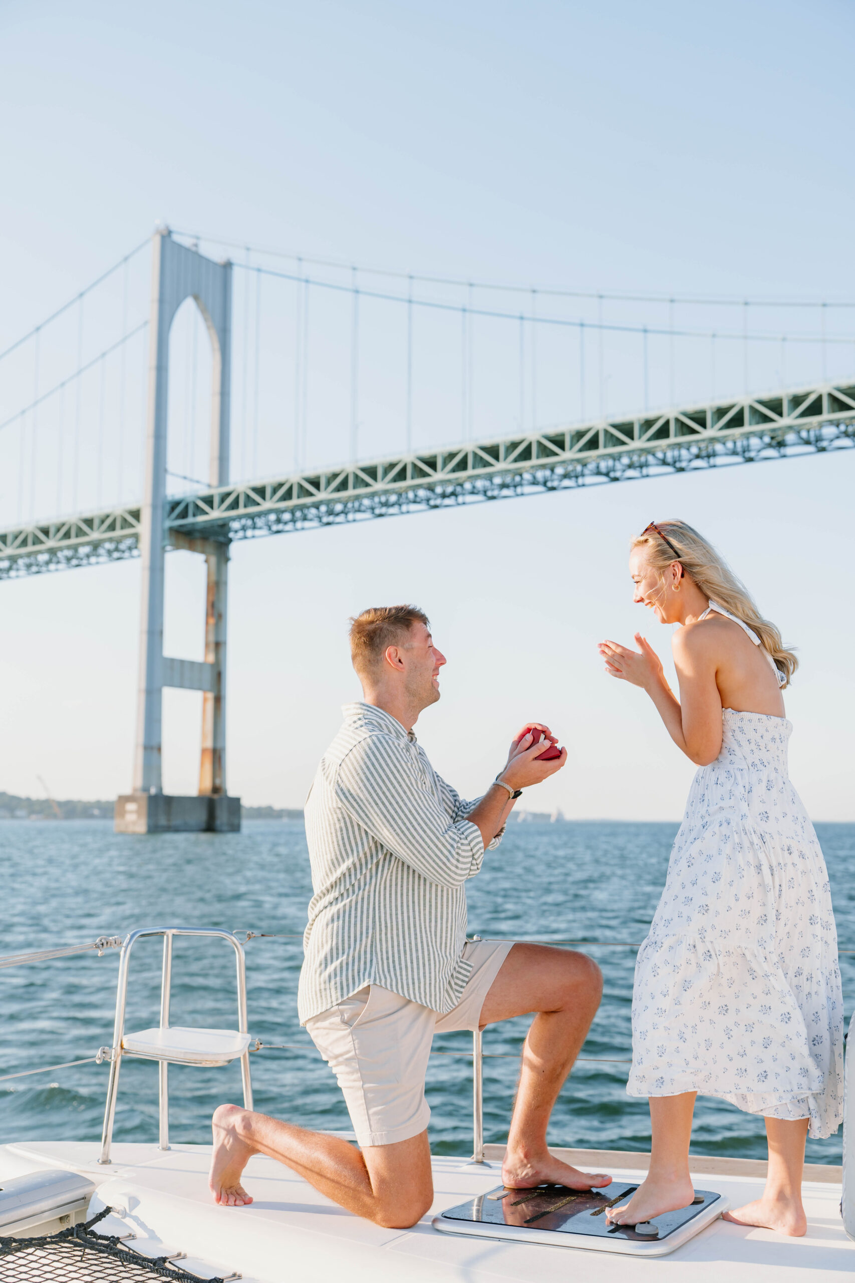 Wedding Photographer Newport Rhode Island | Deirdre Photography Timeless and Organic Moments | A man kneels on a boat, holding a ring box, proposing to a surprised woman in a white dress against the backdrop of a large suspension bridge.