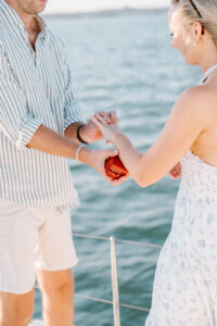 Wedding Photographer Newport Rhode Island | Deirdre Photography Timeless and Organic Moments | A man in a striped shirt holding a ring box proposes to a woman in a floral dress on a boat, with a calm ocean background under soft sunlight.