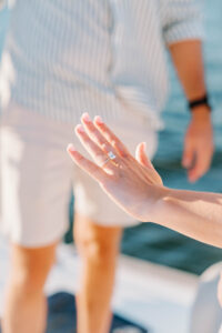 Wedding Photographer Newport Rhode Island | Deirdre Photography Timeless and Organic Moments | A hand with an engagement ring is held up against a blurred background of a person wearing a striped shirt near the water