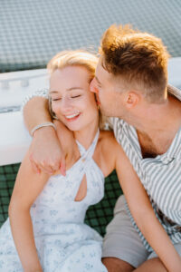 Wedding Photographer Newport Rhode Island | Deirdre Photography Timeless and Organic Moments | A couple sits closely, smiling contentedly. The man in a striped shirt kisses the woman's cheek. She wears a white dress