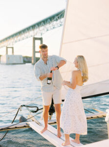 Wedding Photographer Newport Rhode Island | Deirdre Photography Timeless and Organic Moments | A couple stands on a sailboat at sunset near a bridge. The man is opening a champagne bottle, and both appear joyful and relaxed, enjoying the serene water.
